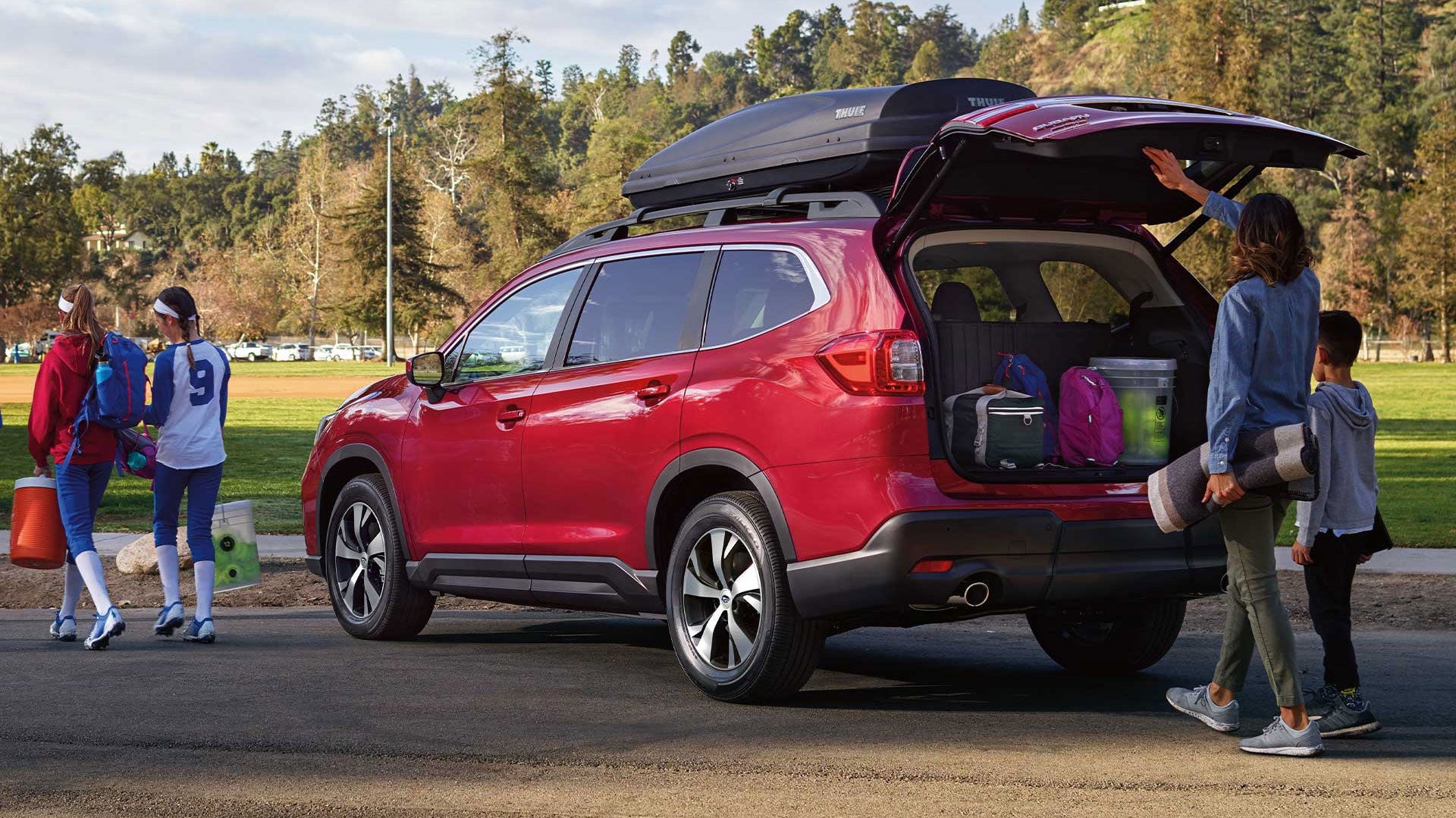 Families packing a red SUV with a roof cargo box for a sports outing, with kids in jerseys nearby.