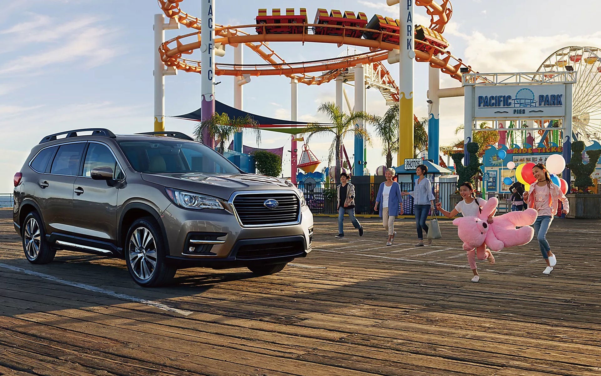 A beige Subaru Ascent SUV parked on a pier with families, and a girl in a pink bunny costume, heading to an amusement park.