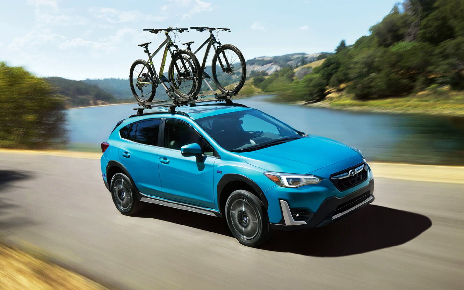 Blue Subaru Crosstrek with two mountain bikes on its roof rack drives along a road next to a lake and hills.