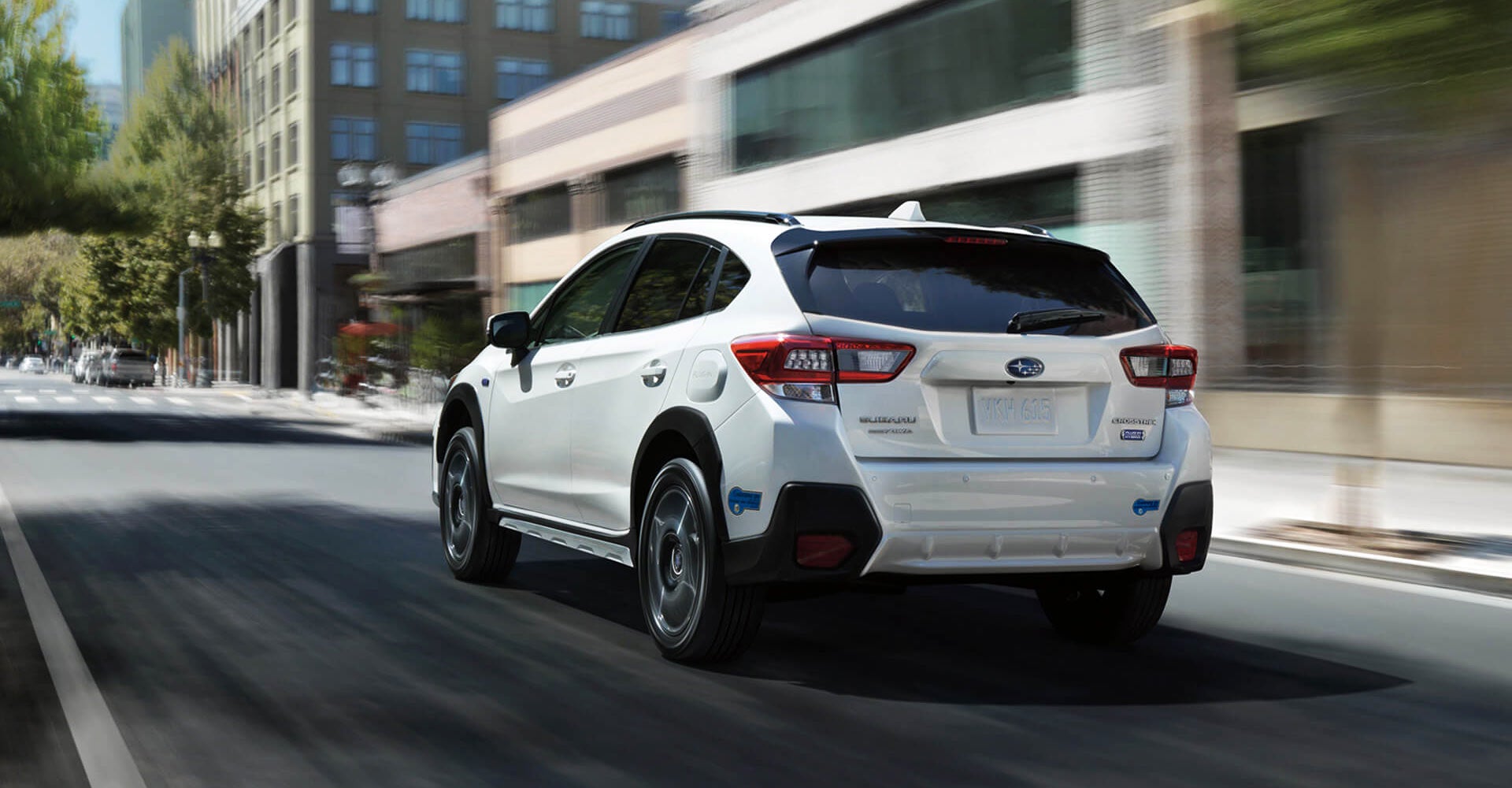 Rear view of a white Subaru Crosstrek hybrid driving down a city street with blurred buildings and trees in the background.