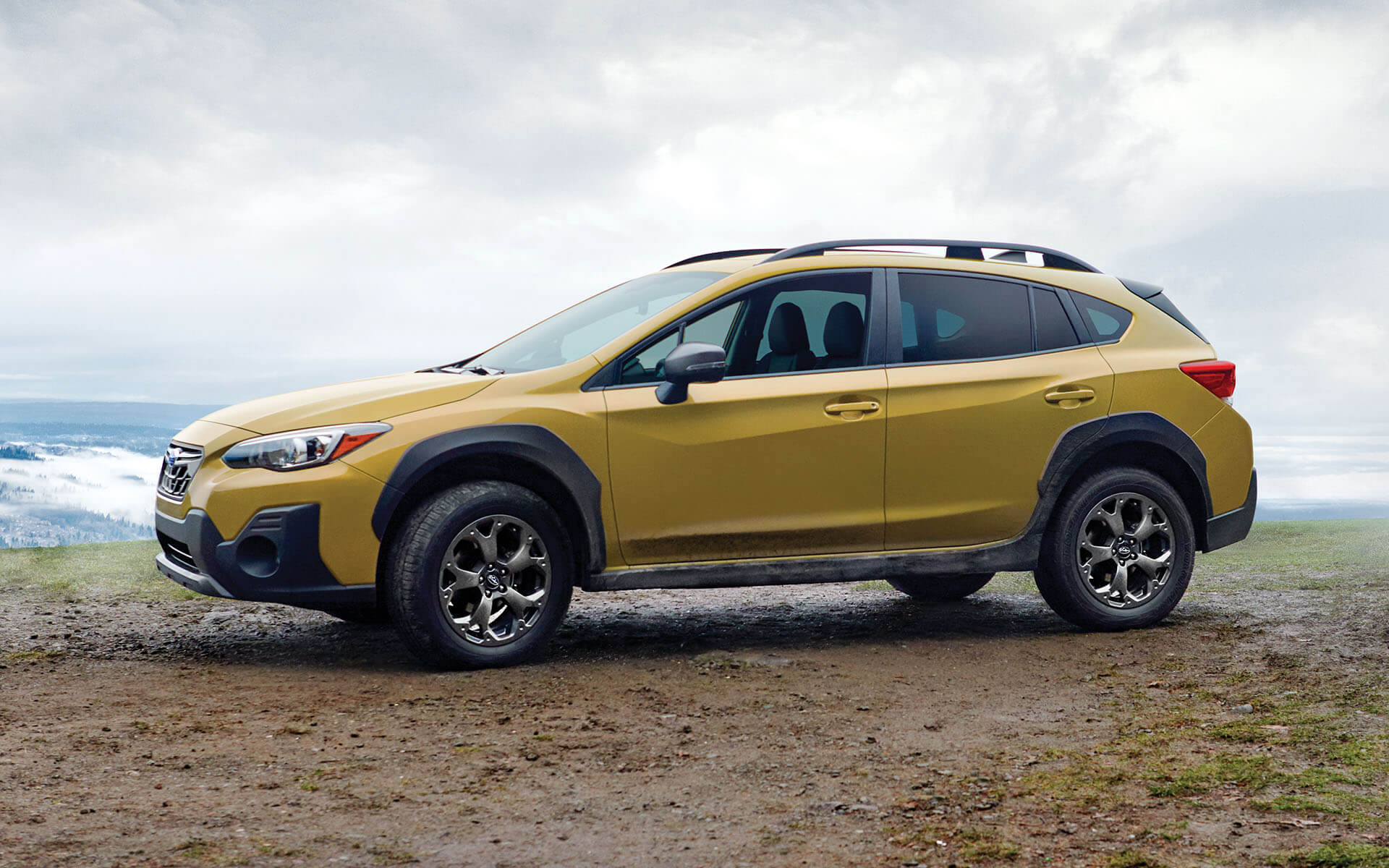 A gold Subaru Crosstrek SUV parked on a dirt road, with a cloudy mountainous landscape in the background.