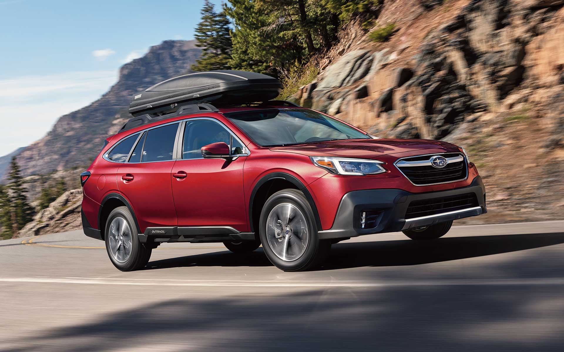 A red Subaru Outback SUV with a black cargo box on its roof, driving quickly on a mountainous road.