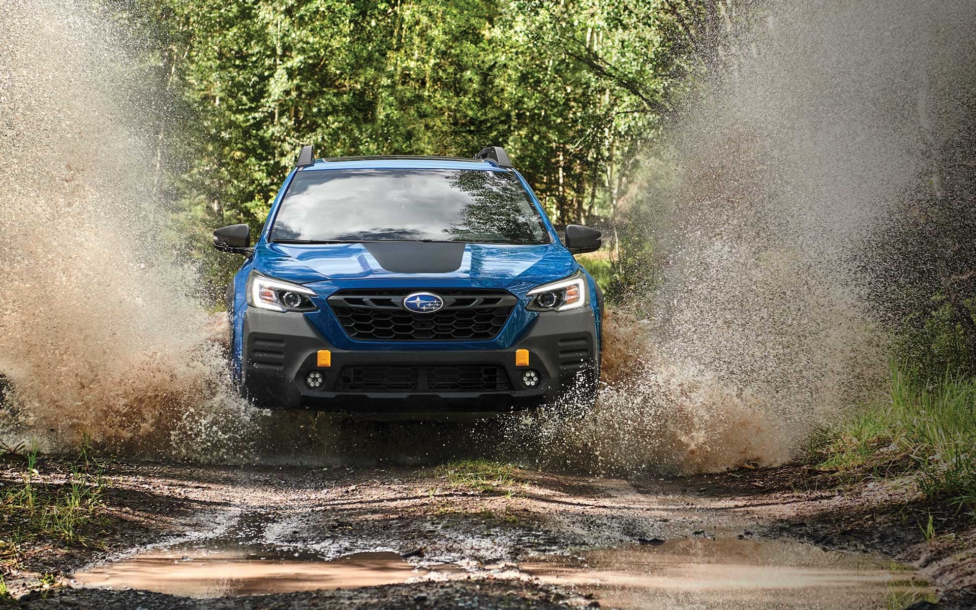 Blue Subaru SUV splashes through a large mud puddle on an off-road trail in a forest.