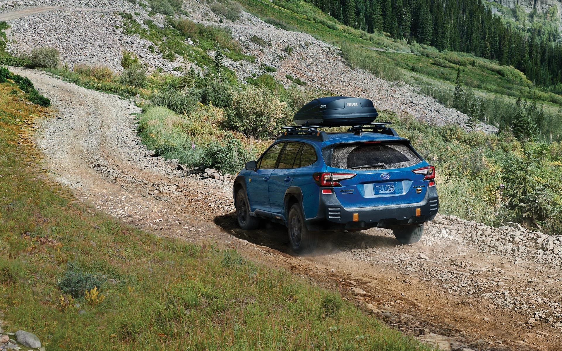 Blue Subaru Outback Wilderness with roof cargo box driving up a dusty mountain trail.