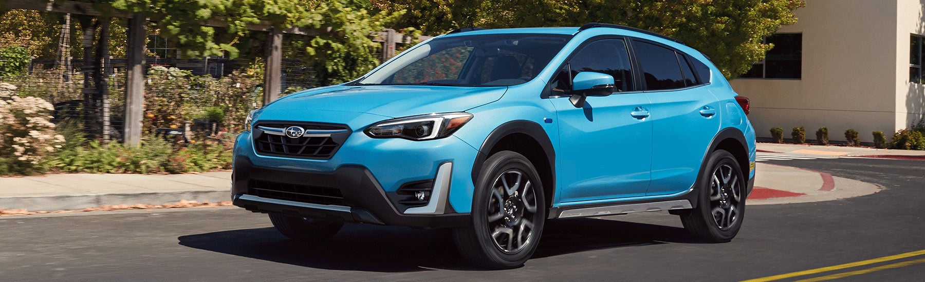 A light blue Subaru Crosstrek with black wheels drives on a curved road lined with green bushes and a building.