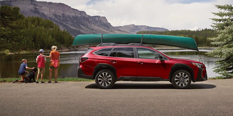Red SUV with a green canoe on its roof rack, parked by a family and dog looking towards a lake and mountains.