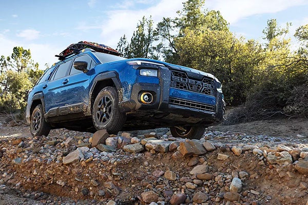 Front three-quarter view of a blue Subaru Outback driving over rugged rocky terrain with a loaded roof basket.