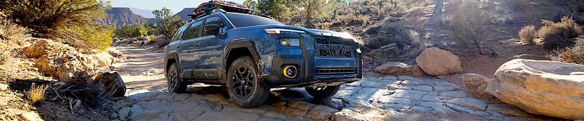 Panoramic shot of a blue Subaru Outback navigating a rocky trail in a desert canyon landscape.