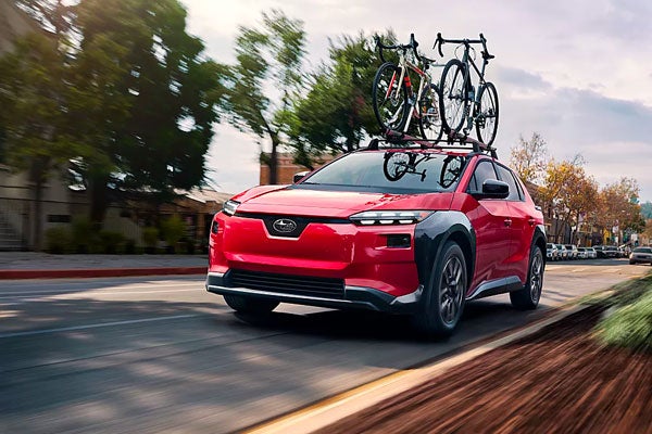 Red Subaru Solterra with two bicycles mounted on its roof rack driving down a city street.
