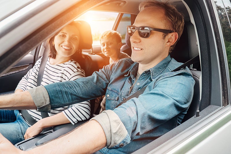 A smiling family, mother and son in the back, father driving, all buckled up in their car.