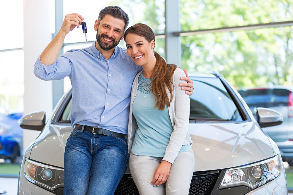 Happy couple posing with their new grey car, man holding up keys, in a bright car dealership showroom.