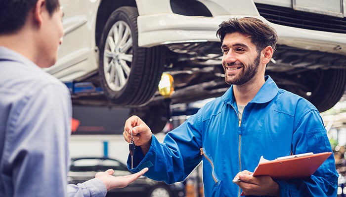 A smiling mechanic in blue overalls hands car keys to a customer